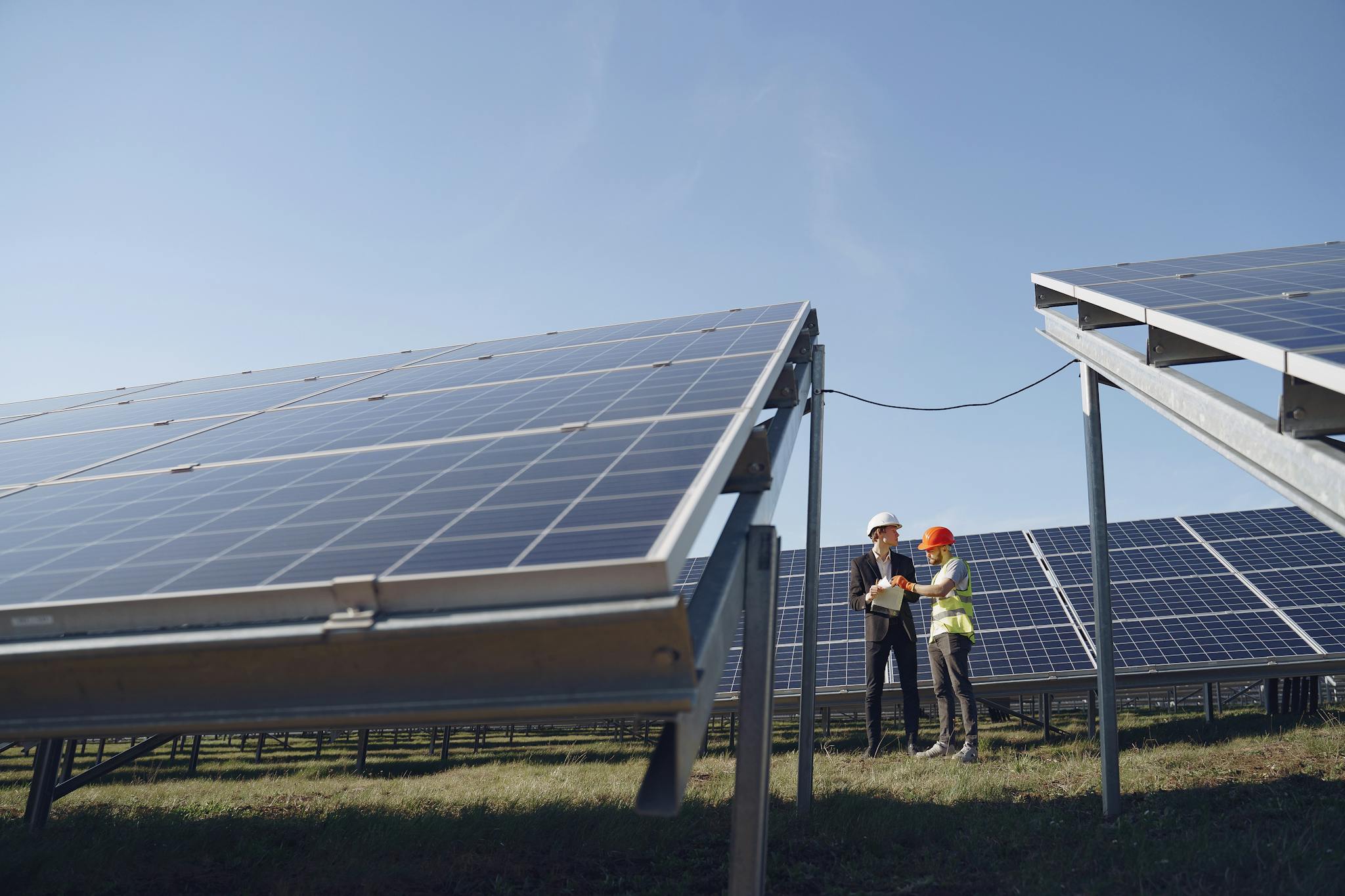 Supervisor in hardhat and formal wear discussing project with workman in uniform while standing with papers near modern solar panels under blue sky